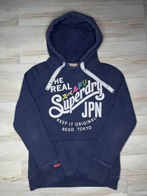Mörkblå Superdry hoodie med tryck - Snygg mörkblå hoodie från Superdry med stor vit logga och färgglada japanska tecken på bröstet🤩Hoodien är i fint skick och passar Xs-S🙌🏻 Hör av er vid funderingar samt snabbaffär😇