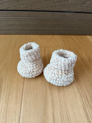 Beige virkade tofflor för baby - Supersöta beige virkade tofflor för baby, gjorda i mjukt och fluffigt garn. De har en rundad form och ribbad kant upptill för extra mysfaktor. Perfekta för att hålla små fötter varma hemma.