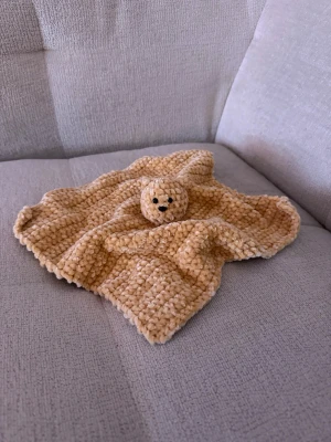 Gosig beige snuttefilt med björnhuvud - Mysig snuttefilt i beige plysch med ett gulligt björnhuvud i mitten. Filten är fyrkantig och har en mjuk, fluffig struktur som känns skön att hålla i. Perfekt som trygghetsskapare eller dekoration i barnrummet. Handgjord