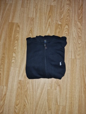 Svart hoodie från Polo Ralph Lauren - Säljer en svart hoodie med dragkedja från Polo Ralph Lauren. Klassisk design med huva, fickor framtill och den ikoniska vita logon broderad på bröstet. Perfekt för en chill och stilren look. Materialet känns mjukt och bekvämt.