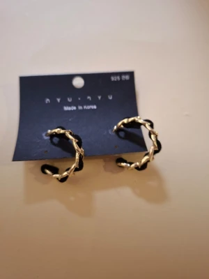 Svart-guldiga hoops  - Säljer ett par snygga örhängen från NYU NYU. De är hoops i en tvinnad design med svart och guldfärgad metall. Örhängena är märkta med 925 och har en modern, trendig look som passar perfekt till din stil.