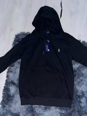 Svart hoodie från Polo Ralph Lauren - Svart hoodie från Polo Ralph Lauren med dragkedja och klassisk huva. Ikonisk vit logga broderad på bröstet och praktiska fickor framtill. Materialet är mjukt och bekvämt, perfekt för en avslappnad och stilren look.