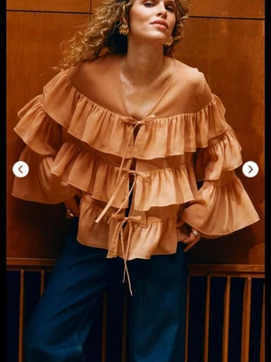 Volangblus i aprikos från Kappahl  - Trendig aprikosfärgad blus  med flera volanglager och knytband framtill. Blusen har offshoulder-modell och vida, långa ärmar. Tillverkad i ett lätt, luftigt tyg som ger en bohemisk och cool vibe.