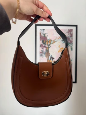 Brun axelremsväska i skinnlook - Snygg brun handväska i halvmåneform med svart axelrem och guldfärgat vridlås. Väskan har en minimalistisk design och är tillverkad i ett material som liknar skinn. Perfekt för dig som gillar stilrena accessoarer.