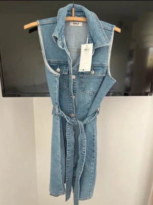 Ärmlös jeansklänning från ONLY - Säljer en snygg ärmlös jeansklänning från ONLY med knappar framtill, bröstfickor och ett matchande bälte i midjan. Klänningen har klassisk krage och är i ljusblå denim, perfekt för en trendig och avslappnad look. Helt ny 