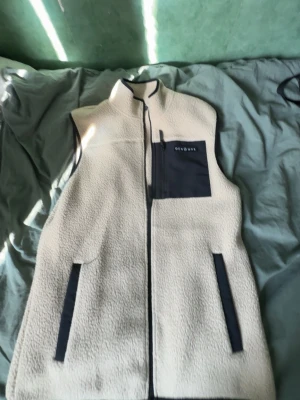 Beige fleeceväst från Dojono - Beige fleeceväst från Dojono med svart dragkedja, hög krage och svarta detaljer. Västen har två sidofickor och en bröstficka med dragkedja och Dojono-logga. Perfekt för lager-på-lager och chill stil. Perfect inför våren