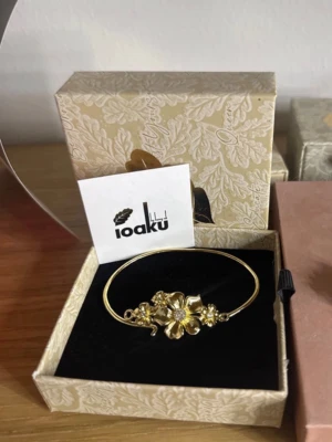 Guldfärgat armband med blommor Ioaku - Elegant guldfärgat armband från Ioaku med en stor blomma i mitten och mindre blommor på sidorna. Armandet är från det trendiga märket IOAKU☺️Den har varit bara en gång i användning och annars i super skick😇Rek. 69,96€. Storleken är 18cm! 
