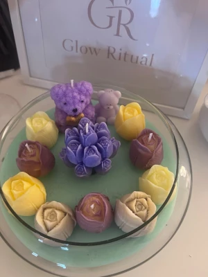 Glow Ritual dekorljus i glasfat - Unikt set med dekorativa ljus från Glow Ritual. Innehåller handgjorda ljus i form av blommor i lila, gult, beige och  samt ett lila björnljus och ett litet ljus i form av en nalle. Allt är placerat på ett runt glasfat med mintgrön botten. Perfekt som dekoration.