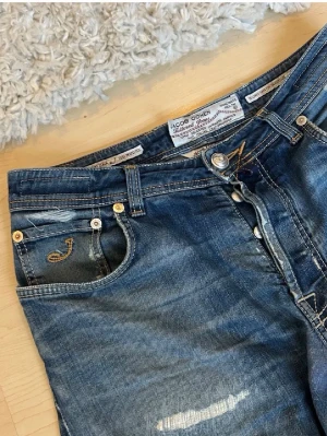 Jacob Cohen jeans - Snygga blå jeans från Jacob Cohen med slitna detaljer och kontrastsömmar. Klassisk femficksmodell med guldfärgade nitar och läderpatch bak i midjan. Jeansen har en broderad logga på fickan och är tillverkade i mjukt denimtyg. Original pris 7000+ Mitt pris 699. Midja: 43 längd: 105 Priset är ej hugget i sten