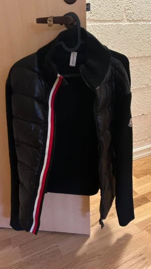 Moncler cardigan s/m - Snygg svart pufferjacka från Moncler med glansig vadderad front och stickade ärmar. Jackan har dragkedja med röd, vit och blå rand samt Moncler-logga på ärmen. Perfekt för kalla dagar och riktigt trendig look.