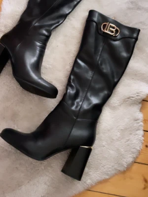 Svarta höga stövlar med klack - Säljer ett par svarta höga stövlar i skinnimitation med chunky klack och guldfärgad detalj upptill. Stövlarna har rundad tå och en dekorativ metallogo på skaftet. Perfekta för att lyfta vilken outfit som helst.