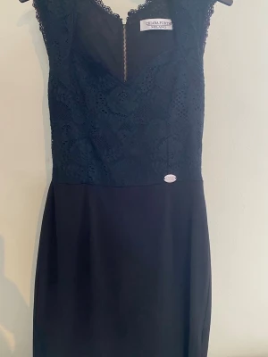 Svart spetsklänning från Chiara Forthi - Elegant svart klänning från Chiara Forthi med spetsdetaljer upptill och v-ringad hals. Klänningen är ärmlös och har en dekorativ dragkedja baktill. Kjoldelen är slät och enkel, vilket ger en stilren look. Perfekt för dig som gillar klassisk och feminin stil.