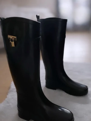 Svarta höga gummistövlar med låsdetalj - Snygga svarta höga gummistövlar med guldfärgad låsdetalj vid skaftet. Stövlarna har en klassisk, stilren form och är tillverkade i blankt gummimaterial. Perfekta för regniga dagar och ger en trendig touch till din outfit.