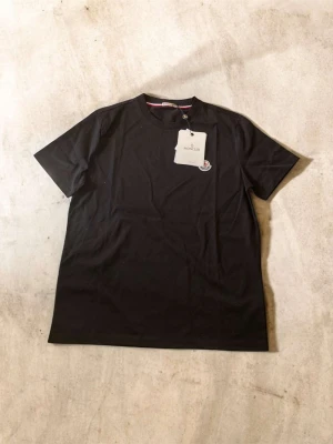 Svart Moncler t-shirt i bomull - Snygg svart t-shirt från Moncler med klassisk rund hals och korta ärmar. Tillverkad i mjuk bomull och har Monclers ikoniska logga på bröstet. Perfekt basplagg med stilren design och diskreta detaljer.