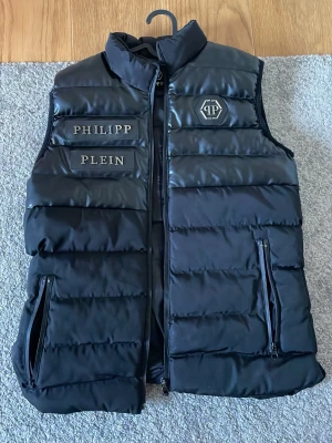 Svart dunväst från Philipp Plein - Snygg svart dunväst från Philipp Plein med stora logoplaceringar på bröstet och en sexkantig PP-logga. Västen har två dragkedjeförsedda fickor och hög krage. Materialet är glansigt och quiltat för en modern look.