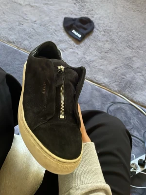 Svarta sneakers från Axel Arigato - Säljer ett par svarta sneakers från Axel Arigato i mocka med vit sula och cool dragkedja istället för snören. Minimalistisk design med diskret logga på sidan. Perfekta för dig som gillar stilrena och moderna skor.