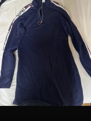 Marinblå långärmad klänning - Sportig marinblå klänning med lång ärm och hög krage. Dragkedja framtill med ringdetalj och vita ränder med text längs ärmarna. Perfekt för en avslappnad och trendig look.