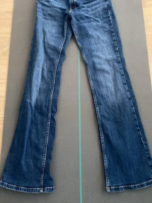 Blå bootcut jeans med slitningar - Säljer ett par blå bootcut jeans med lätt slitna detaljer framtill. Jeansen har klassisk femficksmodell och normal midja. Materialet är denim i bomull och passformen är utsvängd nedtill för en retro vibe.