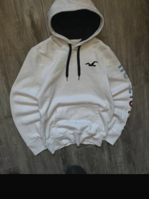 Vit hoodie från Hollister - Säljer en vit hoodie från Hollister med svart logga på bröstet och svarta snören i huvan. Hoodien har känguruficka och tryck i blått och rött på ena ärmen. Insidan av huvan är svart och materialet känns mjukt och skönt.