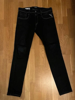 Svarta anbass jeans från Replay - Säljer ett par svarta slim fit jeans från Replay med vita kontrastsömmar och klassiska fem fickor. Jeansen har en snygg, smal passform och detaljer som Replay-logga på bakfickan och läderpatch i midjan. Perfekta för en trendig och stilren look.