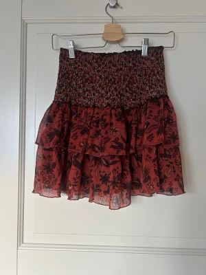 Röd volangkjol med blommigt mönster - Supersöt röd kortkjol med svarta blommor och volanger. Kjolen har smock i midjan för en bekväm passform och är tillverkad i ett lätt, luftigt material. Perfekt för att skapa en trendig och avslappnad look.