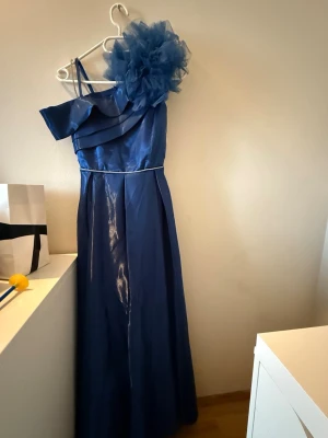 Marinblå långklänning oneshoulder - Snygg marinblå långklänning med oneshoulder-design och stor volangdetalj i tyll på axeln. Klänningen har en elegant siluett och tunna axelband på ena sidan. Perfekt för dig som vill sticka ut på bal eller fest.