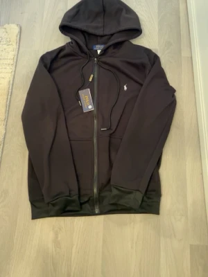 Svart hoodie - Snygg svart hoodie med klassisk design med huva och dragsnören, samt den ikoniska vita logotypen broderad på bröstet. Tillverkad i mjukt bomullsmaterial och har fickor framtill. Perfekt för en avslappnad och stilren look.