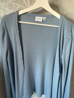Ljusblå ribbad kofta från Vila - Ljusblå kofta från Vila med ribbad struktur och långa ärmar. Koftan har öppen front och fina vågiga kanter längs öppningen. 