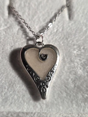 Glow in the dark halsband med hjärthänge - Unikt halsband med ett hjärtformat hänge i silverfärgad metall och vit glittrig insida som lyser starkt blått i mörkret. Hänget har dekorativa snirkliga detaljer och kedjan är tunn och silverfärgad i rostfritt stål. Perfekt för dig som gillar smycken med en twist och vill sticka ut.