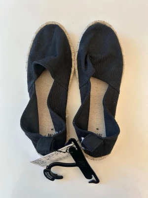 Svarta espadrillesandaler  - Säljer ett par svarta espadrillesandaler med ovandel i tyg och klassisk flätad sula i jute. Skorna har en enkel slip-in design och rundad tå. Perfekta för sommaren och lätta att matcha med olika outfits, med prislapp kvar 