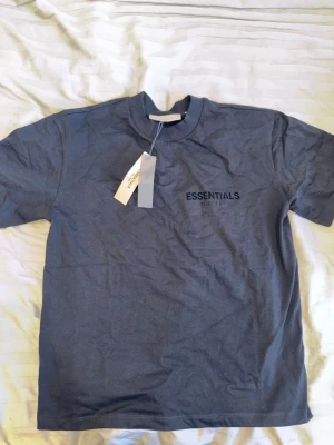Svart Essentials T-shirt - Snygg svart t-shirt från Fear of God Essentials med tryck både fram och bak. Klassisk passform med rund hals och korta ärmar. Perfekt för dig som gillar streetwear och vill ha en stilren look.