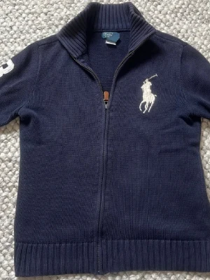 Mörkblå ziptröja från Polo Ralph Lauren - Mörkblå stickad ziptröja från Polo Ralph Lauren med hög krage och vit broderad logga på bröstet. Tröjan har ribbade muddar och dragkedja framtill. Snygg detalj med siffra på ärmen.