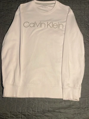 Vit långärmad tröja Calvin Klein - Den är storlek M men passar S också för den är slim fit. Vit långärmad tröja från Calvin Klein med rund halsringning och logga broderad framtill. Tröjan är i mjuk bomull och har ribbade muddar vid ärmslut och nederkant. Enkel och stilren design som passar till det mesta.