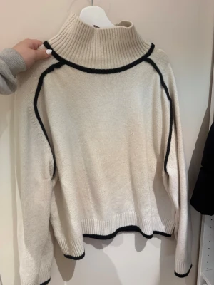 Beige tröja med svarta detaljer H&M - Stickad beige tröja från H&M med svarta kontrastlinjer längs axlar, ärmar och kanter. Tröjan har hög ribbad krage, långa ärmar och avslappnad passform. Perfekt för lager-på-lager och snygg till jeans eller kjol.