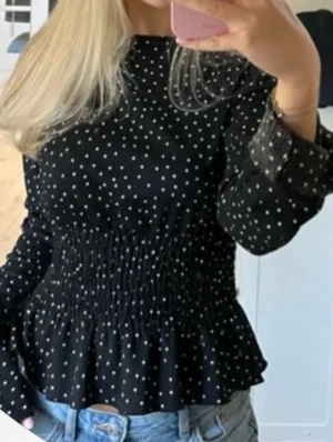 Svart prickig blus med smock - Svart blus med vita prickar och smockad midja som ger en snygg siluett. Långa ärmar med lätt volang vid ärmslut och en liten volang nertill. 