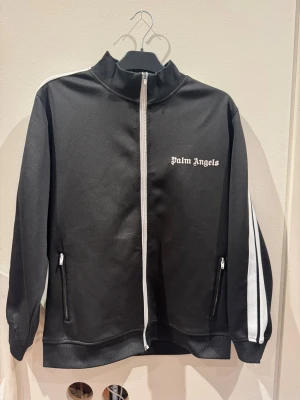 Svart track jacket från Palm Angels - Säljer en svart track jacket från Palm Angels med vita ränder längs ärmarna och vit dragkedja. Jackan har hög krage, två fickor med dragkedja och logga på bröstet. Perfekt för en sportig och trendig look.