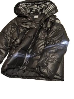 Svart dunjacka från Burberry - Säljer en svart dunjacka från Burberry med klassiskt rutigt foder i huvan och tryck på ärmen. Jackan har dragkedja, knappar och en stor huva. Perfekt för kalla vinterdagar och riktigt snygg streetstil.