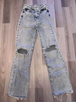 Ljusblå slitna raka jeans - Säljer ett par ljusblå raka jeans med hög midja och stora slitningar vid knäna. Jeansen har fransiga benslut och klassisk femficksdesign. Perfekta för en avslappnad och trendig streetstil.