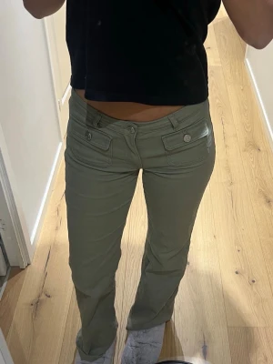 Olivgröna jeans  med framfickor - Säljer ett par olivgröna jeans med raka ben och låg midja. Jeansen har två stora fickor framtill med knappdetaljer som ger en cool look. Tillverkade i ett mjukt material som känns bekvämt hela dagen.