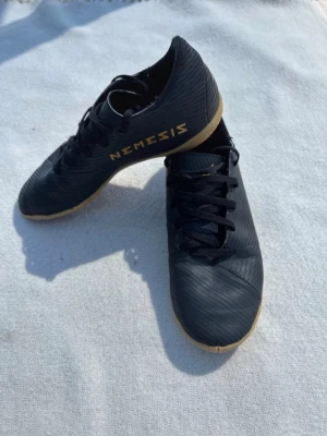 Adidas Nemesis svarta fotbollsskor - Säljer ett par svarta Adidas Nemesis fotbollsskor med gummisula i beige. Skorna har snörning och är tillverkade i syntetmaterial med diskret mönster och guldtext på sidan. Perfekta för inomhusspel och snabba vändningar på planen.