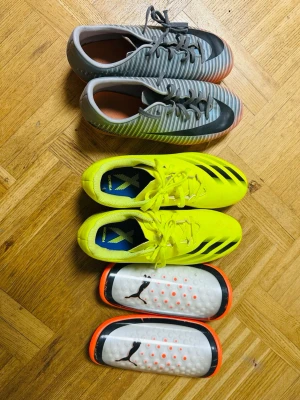 Adidas X Ghosted.4 och Nike Mercurial - Två par fotbollsskor: ett par Adidas X Ghosted.4 i neon-gult med svarta detaljer och ett par Nike Mercurial i grått med svarta och orange detaljer. Båda paren har snörning och låg profil. Medföljer vita benskydd från Puma med orange detaljer. Perfekt för dig som vill sticka ut på planen. Allt för 400kr