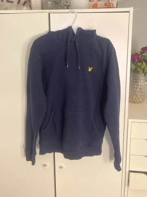 Mörkblå hoodie från Lyle & Scott - Mörkblå hoodie från Lyle & Scott med klassisk gul logga på bröstet. Tröjan har huva med dragsko och en stor magficka framtill. för en chill och avslappnad stil. Materialet känns mjukt och skönt mot huden.