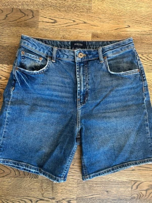 Blå jeansshorts från Pieces - Klassiska blå jeansshorts från Pieces med fem fickor, knapp och dragkedja framtill. Normal midja. Shortsens denim är i bomull och har en rak passform. Perfekta för varma dagar och enkel att matcha med olika toppar.