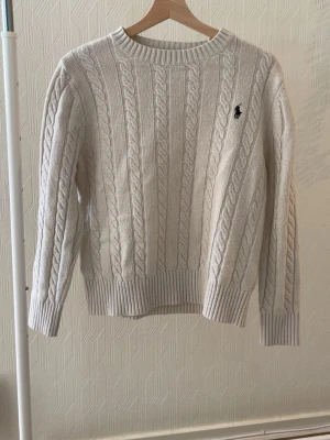 Kabelstickad tröja från Ralph Lauren - Kabelstickad offwhite tröja från Ralph Lauren med rund halsringning och klassisk svart logga broderad på bröstet. Tröjan har långa ärmar och breda ribbade muddar vid ärmslut och nederkant. Perfekt för lager-på-lager och stilrena outfits.
