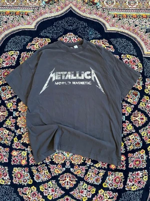 Metallica World Magnetic t-shirt - Svart t-shirt med Metallica World Magnetic-tryck på bröstet och turnédatum på ryggen. Klassisk passform med rund hals och korta ärmar. Perfekt för dig som älskar bandmerch och vill sticka ut med en ikonisk look.