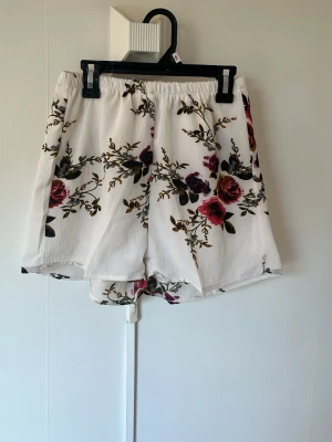 Vita blommiga shorts med resårmidja - Säljer ett par vita shorts med blommigt mönster. Shortsen har en avslappnad passform och elastisk midja för extra komfort. Perfekta för varma dagar och ger en fräsch och somrig look. Inget märke.