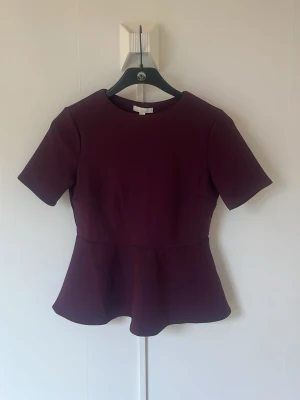 Mörklila peplum topp med kort ärm - Snygg mörklila peplum topp med rund halsringning och korta ärmar. Toppen har en markerad midja och utsvängd nederdel som ger en feminin siluett. Perfekt att styla med jeans eller kjol för en trendig look.