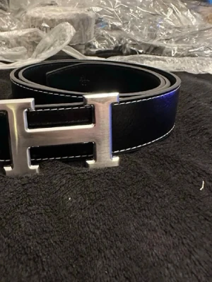 Svart läderbälte H bälte - Snyggt svart bälte i läder med ett stort, silverfärgat H-format metallspänne. Bältet har vita kontrastsömmar längs kanterna som ger en stilren look. Perfekt accessoar för att lyfta vilken outfit som helst.