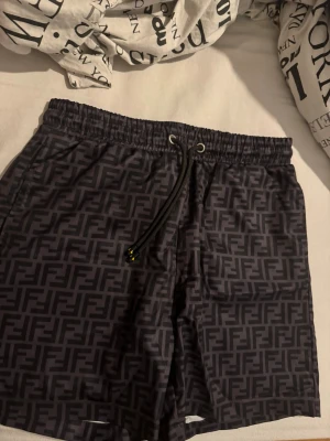 Svarta Fendi badbyxor med logomönster - Snygga svarta badbyxor från Fendi med klassiskt FF-logomönster över hela tyget. De har elastisk midja med snörning och meshfoder på insidan. Perfekta för strand eller pool och tillverkade i lätt syntetmaterial. Fu kar även för daglig användning 