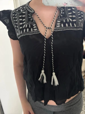 Svart blus med broderi och knyt - Svart blus med vita broderade detaljer på axlar och bröst, samt dekorativa knytband med tofsar framtill. Blusen har korta volangärmar och v-ringning. Perfekt för sommaren. Säljer då den inte kommer till användning
